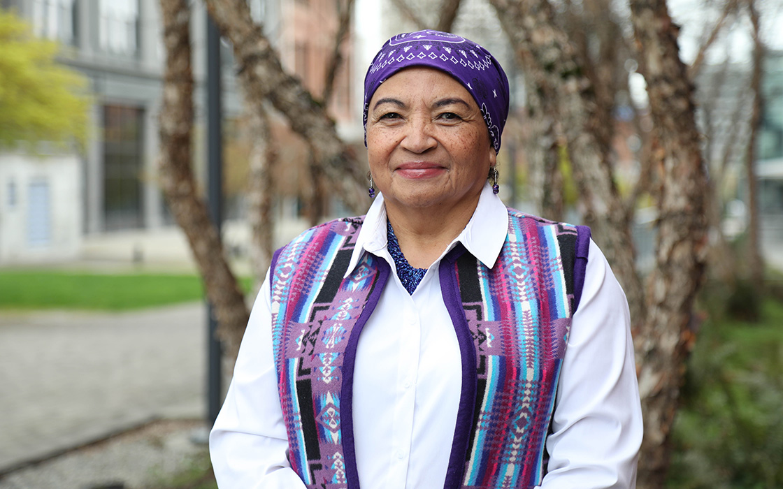 UW Tacoma Ed.D. student Ada McDaniel stands in front of a tree. She is wearing a head wrap that is purple and has symbols woven into it. McDaniel is also wearing a purple vest with symbols woven into it. She has on a white undershirt.
