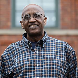 Headshot of UW Tacoma staff member Jamal Gabobe. Gabobe is wearing a red plaid shirt and has on glasses. A brick building with a window is in the background.