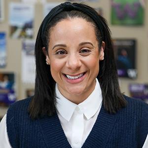 Headshot of staff member Navia Winderling. Navia has long, dark hair. She is wearing a purple sweater vest over a white shirt. There are a handful of UW Tacoma brochures hanging from the wall behind her.