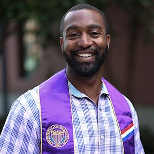 Headshot of UW Tacoma alumnus Joe Davis. Davis is wearing a blue plaid shirt with a purple stole around his neck that says "veteran." He has short black hair and a beard.